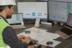 Male engineer wearing hardhat n office and using computer designing blueprints